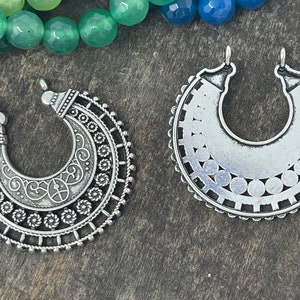 May include: Two silver metal pendant charms in the shape of a crescent moon. One charm has a detailed design with swirls and circles, while the other has a more simple design with a circle pattern.