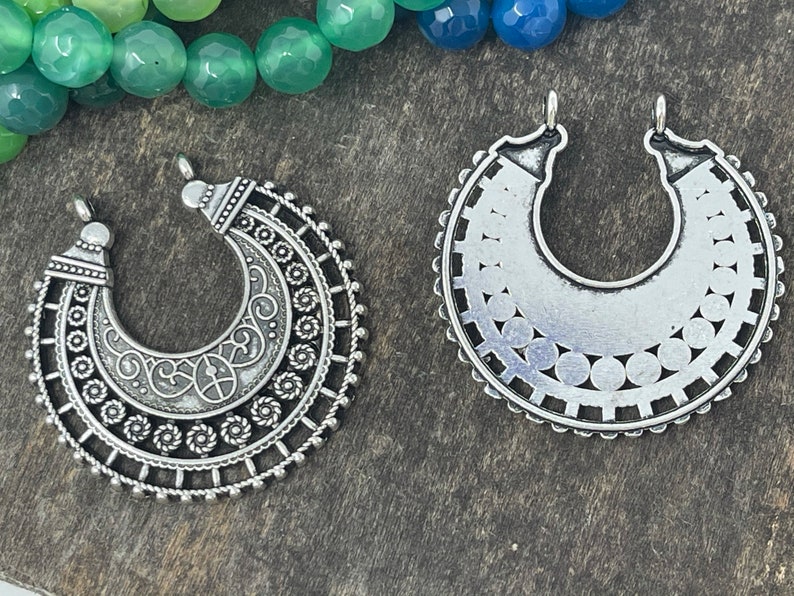 May include: Two silver metal pendant charms in the shape of a crescent moon. One charm has a detailed design with swirls and circles, while the other has a more simple design with a circle pattern.