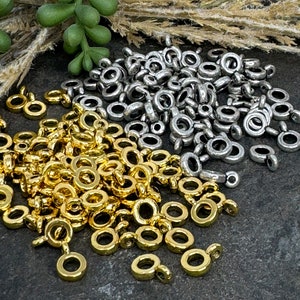 May include: A collection of small metal jump ring connectors with attached loops, featuring a mix of shiny gold-tone and matte silver-tone finishes. These circular jewelry findings are scattered in two distinct piles on a dark textured surface.