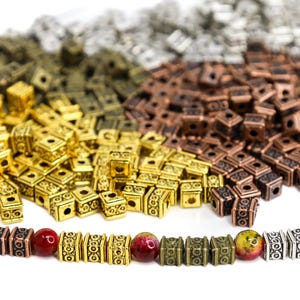 Puede incluir: Un surtido de cuentas cuadradas de metal en tonos dorados, bronce, plateado y cobrizo. Algunas cuentas están ensartadas con cuentas redondas rojas y amarillo-verdosas, para la creación de joyas.