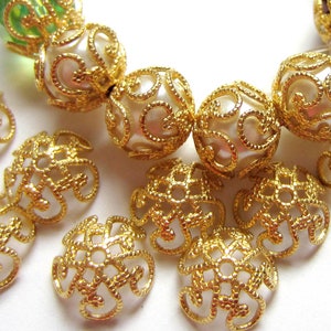 May include: Close-up of various decorative beads. The beads include gold filigree caps and round beads with a pearl-like center and gold filigree overlay. Other beads are green and brown with gold accents. The beads are for jewelry making.