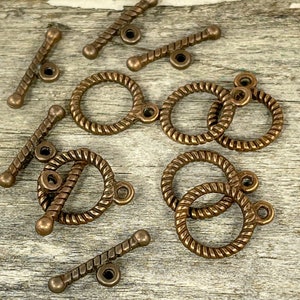 May include: A collection of antique brass toggle clasps and jump rings. The clasps are rope-like and have a round ring on one end and a bar with a round ring on the other end. The jump rings are round and have a rope-like texture.