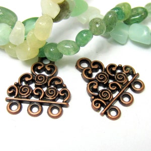 May include: Two ornate copper-colored jewelry clasps with a decorative swirl design. The clasps have multiple loops for attaching to jewelry. In the background, there are strands of colorful gemstone beads in shades of green, yellow, and blue.