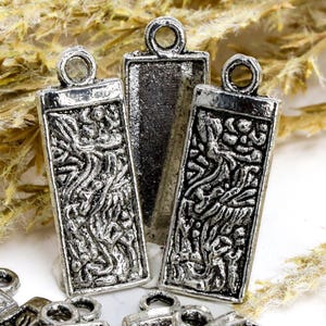 May include: Close-up of several silver-colored rectangular pendants with a loop at the top. Two pendants have an ornate, textured design, while the center one is plain. The pendants are likely for jewelry making or crafting.