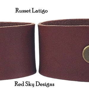 K&ouml;nnte beinhalten: Zwei Lederarmb&auml;nder, eines mit einem Druckknopfverschluss und eines ohne. Beide sind in einem satten Rotbraun gehalten. Die Armb&auml;nder sind aus Leder gefertigt und haben einen rustikalen Look. Der Text "Russet Latigo" und "Red Sky Designs" ist auf dem Bild zu sehen.
