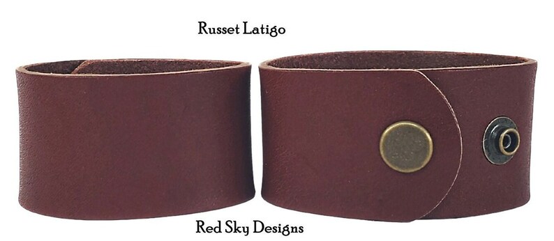 K&ouml;nnte beinhalten: Zwei Lederarmb&auml;nder, eines mit einem Druckknopfverschluss und eines ohne. Beide sind in einem satten Rotbraun gehalten. Die Armb&auml;nder sind aus Leder gefertigt und haben einen rustikalen Look. Der Text "Russet Latigo" und "Red Sky Designs" ist auf dem Bild zu sehen.