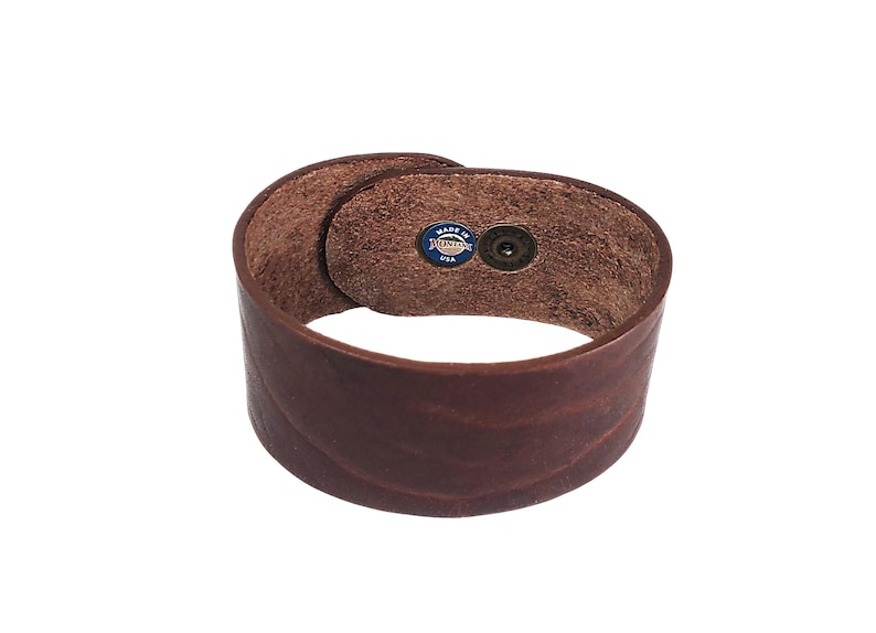 K&ouml;nnte beinhalten: Ein braunes Lederarmband mit einem Druckknopfverschluss. Das Armband hat ein "Made in USA"-Etikett auf der Innenseite.