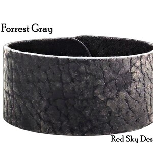 K&ouml;nnte beinhalten: Ein breites, schwarzes Lederarmband mit einer strukturierten, rissigen Oberfl&auml;che. Das Armband ist mit "Forrest Gray" und "Red Sky Designs" beschriftet.