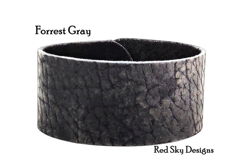 K&ouml;nnte beinhalten: Ein breites, schwarzes Lederarmband mit einer strukturierten, rissigen Oberfl&auml;che. Das Armband ist mit "Forrest Gray" und "Red Sky Designs" beschriftet.