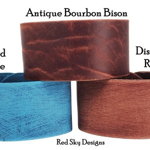 K&ouml;nnte beinhalten: Drei Lederarmb&auml;nder in verschiedenen Farben und Texturen. Das obere Armband ist braun mit einem abgenutzten Look und ist mit "Antique Bourbon Bison" beschriftet. Das mittlere Armband ist blau mit einem abgenutzten Look und ist mit "Distressed Turquoise" beschriftet. Das untere Armband ist rotbraun mit einem abgenutzten Look und ist mit "Distressed Russet" beschriftet.