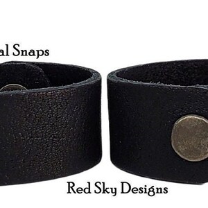 K&ouml;nnte beinhalten: Zwei schwarze Lederarmb&auml;nder mit Schnappverschl&uuml;ssen aus Metall. Die Armb&auml;nder sind von der Seite gezeigt, wobei die Schnappverschl&uuml;sse sichtbar sind. Der Text "Pure Black" und "Shown With Gun Metal Snaps" ist &uuml;ber den Armb&auml;ndern sichtbar. Der Text "Red Sky Designs" ist unter den Armb&auml;ndern sichtbar.