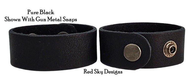 K&ouml;nnte beinhalten: Zwei schwarze Lederarmb&auml;nder mit Schnappverschl&uuml;ssen aus Metall. Die Armb&auml;nder sind von der Seite gezeigt, wobei die Schnappverschl&uuml;sse sichtbar sind. Der Text "Pure Black" und "Shown With Gun Metal Snaps" ist &uuml;ber den Armb&auml;ndern sichtbar. Der Text "Red Sky Designs" ist unter den Armb&auml;ndern sichtbar.