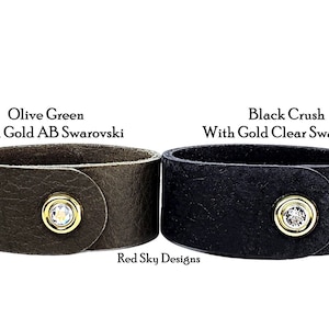 May include: Two leather bracelets with gold snap closures. The bracelet on the left is olive green with a gold AB Swarovski crystal. The bracelet on the right is black with a gold clear Swarovski crystal. Both bracelets have the text "Red Sky Designs" printed below them.