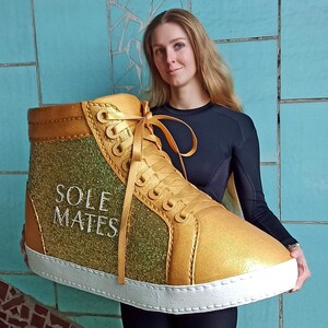 May include: A large gold and green glitter high-top trainer with white laces and a white sole. The shoe has the words "SOLE MATES" written on the side.