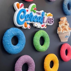 May include: A black wall with a white sign that says "The Cereal Lab" in blue and orange letters. There are several colorful donut-shaped decorations on the wall, including green, pink, purple, blue, and yellow.