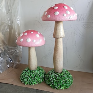 Könnte beinhalten: Zwei rosa-weiße Pilz-Dekorationen mit weißen Punkten. Die Pilze sind aus einem strukturierten Material gefertigt und haben ein realistisches Aussehen. Jeder Pilz sitzt auf einem Bett aus grünem Laub.
