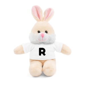 May include: A plush bunny toy with a white t-shirt featuring a large black letter "R". The bunny has cream-colored fur, pink inner ears, and a pink nose. The toy is designed to be soft and cuddly.