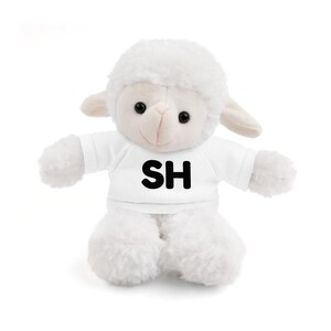 May include: A white plush lamb toy with a fluffy body, head, and limbs. The lamb wears a white t-shirt with the black letters "SH" printed on the front. The lamb has black eyes and a pink nose.