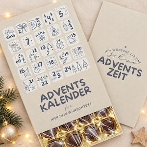 May include: A beige advent calendar with dark blue text and illustrations for numbers one through twenty-four. The text reads ADVENTS KALENDER, HIER DEIN WUNSCHTEXT, and ICH WÜNSCHE DIR EINE SCHÖNE ADVENTS ZEIT. A tray of round chocolates is visible at the bottom.