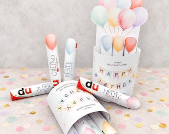 Può includere: Una collezione di tubi cilindrici bianchi per bomboniere e un contenitore abbinato, decorati con illustrazioni di palloncini ad acquerello e festoni pastello. Il testo include frasi come Happy Birthday, du bleibe e Beste.