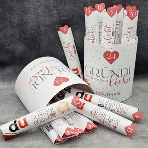 Könnte beinhalten: Ein weißer Behälter mit roten Herzen und dem Text "GRÜNDE WARUM ICH DICH LIEBE" enthält aufgerollte Papierrollen. Jede Rolle hat eine Nummer und eine Nachricht. Einige Rollen sind um den Behälter herum verstreut. Das Wort "du" ist sichtbar.