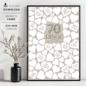 May include: A framed print with a beige background and numerous white heart shapes. The print features the text "70 Dinge die wir dir w&uuml;nschen" in the center, with handwritten wishes inside the hearts. A black vase with a plant is in the foreground.