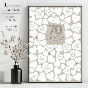 May include: A framed print with a beige background filled with white heart shapes. The print has the text "70 Things We Wish" in the center, with handwritten wishes inside the hearts. A black vase with a plant is in the foreground.