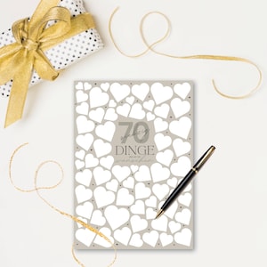 May include: A greeting card with a gray background covered in white hearts. The card has the number 70 and text in German. A black and gold pen rests on the card, with a wrapped gift and gold ribbon in the background.