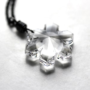 May include: A clear crystal snowflake pendant on a black cord necklace. The snowflake is faceted and has a starburst design.