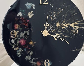 May include: A round black wall clock with gold numerals and hands. The clock face features dried flowers and gold accents. The clock is displayed on a white stand. The clock's diameter is approximately 10 inches.