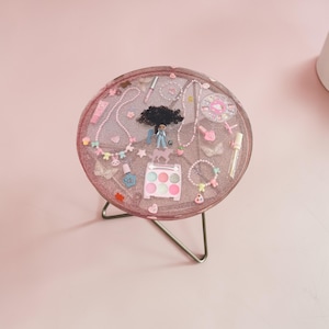 May include: A circular, pink glitter resin table with a metal frame. The table is adorned with miniature jewellery, a doll, and makeup. Items include necklaces, bracelets, nail polish, and a makeup palette. The table is set against a pink backdrop.