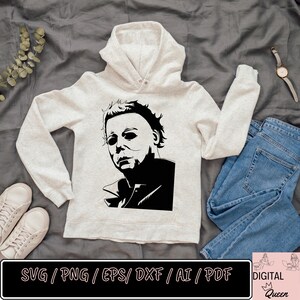 May include: A white hoodie with a black and white illustration of Michael Myers from the Halloween horror movie franchise. The image is a silhouette of Michael Myers's face and upper body. The hoodie is on a gray surface with a pair of white sneakers, blue jeans, and a watch.