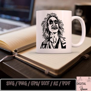 May include: A white ceramic mug with a black and white illustration of a character from the movie "Beetlejuice". The character is shown with a wide open mouth and a menacing expression. The mug is sitting on a wooden table with a notebook and a pen.