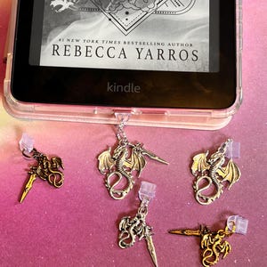 May include: An e-reader in a clear case shows the cover of "Storm" by Rebecca Yarros. Several dragon-shaped charms with swords are attached to the e-reader. The charms are silver and gold.
