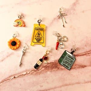 May include: A collection of small charms, including a rainbow, sunflower, caution sign, bow, chili pepper with handcuffs, a book with "Book Worm" text, a wine bottle with a glass, and a silver arrow. The charms are attached to clear clips.