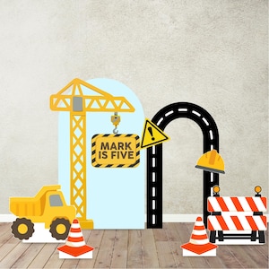 May include: A construction-themed birthday party decoration set. Includes a yellow dump truck, crane, road, caution signs, and a sign that reads "MARK IS FIVE". The set also features orange and white traffic cones and a construction barrier.