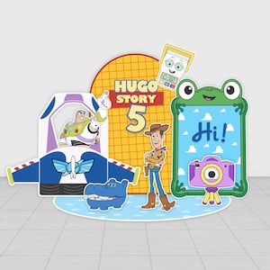 May include: A decorative party display featuring a yellow grid background with the text Hugo Story 5, a Buzz Lightyear cutout, a Woody cutout, a blue hippo, a green frog-shaped frame with the text Hi!, a purple camera, and a small calculator.