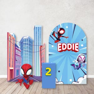 May include: A blue and red cardboard backdrop with a cityscape and two cartoon Spider-Man characters. The backdrop is decorated with stars and the name "EDDIE" in white letters. The backdrop is numbered "2".