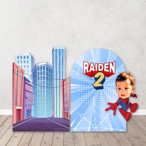 May include: A birthday party decoration set featuring a cityscape backdrop and a Spider-Man-themed arch. The arch displays the name "RAIDEN" and the number "2", with a cartoon image of a child dressed as Spider-Man.