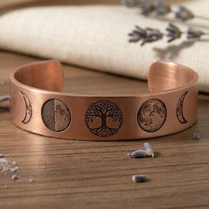 May include: A copper-colored cuff bracelet featuring engraved phases of the moon and a tree of life design. The bracelet is open-ended and rests on a wooden surface, with sprigs of lavender in the background.