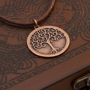 May include: A copper-colored pendant necklace featuring a tree of life design. The pendant hangs on a braided brown leather cord. The necklace rests on a dark brown wooden box with ornate carvings and a metal clasp.