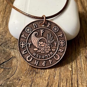 May include: A copper-colored pendant necklace with a brown leather cord. The round pendant features a detailed design of a cornucopia, wheat, and runes. The necklace is displayed on a white surface against a wooden background.