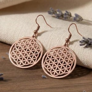 Puede incluir: Pendientes de oro rosa con un diseño geométrico circular. Los pendientes se muestran sobre una superficie de madera, con ramitas de lavanda y un paño de color crema en el fondo.