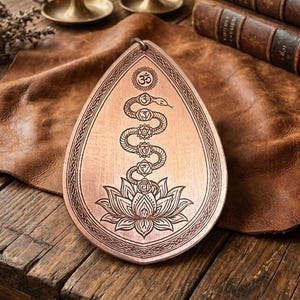 May include: Copper-coloured, teardrop-shaped wall hanging with an engraved design. The design includes a snake, lotus flower, and chakra symbols. The wall hanging is displayed on a wooden table with candles and books in the background.