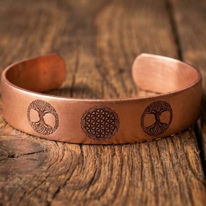 Puede incluir: Una pulsera de brazalete de cobre con tres diseños grabados: un árbol de la vida, una flor de la vida y otro árbol de la vida. La pulsera se muestra sobre una superficie de madera rústica, mostrando sus cálidos tonos metálicos.