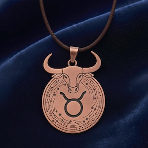 May include: A copper-coloured pendant necklace featuring the Taurus zodiac symbol. The pendant has a bull's head design above the Taurus symbol, with a brown cord. The background is a dark blue fabric.