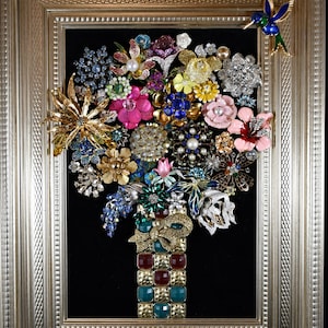 Puede incluir: Una obra de arte enmarcada con un ramo de broches de flores coloridas. Los broches están dispuestos sobre un fondo negro dentro de un marco dorado. Un broche de colibrí está adherido al marco. El ramo incluye broches dorados, rosas, azules y plateados.