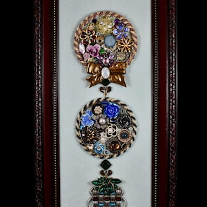 Può includere: Opera d'arte incorniciata con due composizioni circolari di gioielli e abbellimenti vintage, tra cui fiori, perline e bottoni. Un fiocco dorato e un elemento decorativo inferiore completano il design. La cornice è marrone scuro.