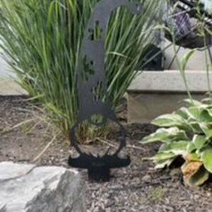 May include: Black metal garden gnome silhouette with a whimsical design. The gnome is standing on a round base and has a long, pointed hat with a butterfly design. The gnome is surrounded by green grass and a rock.