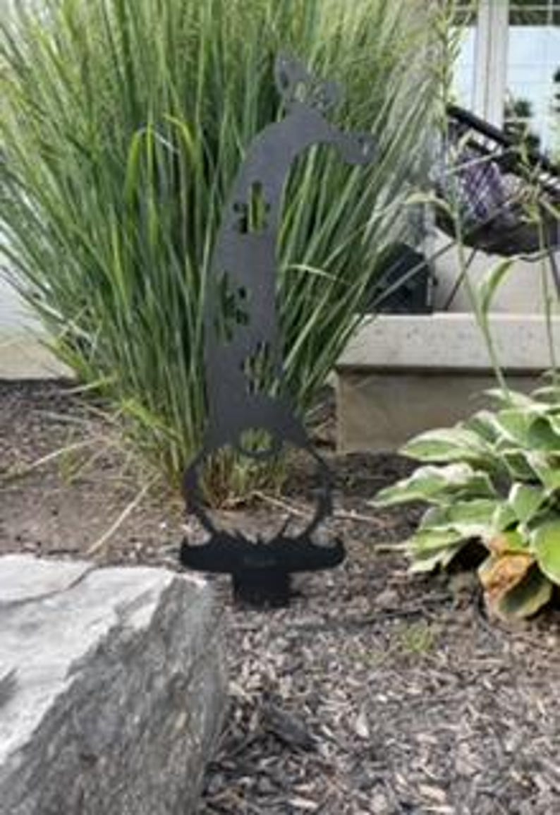 May include: Black metal garden gnome silhouette with a whimsical design. The gnome is standing on a round base and has a long, pointed hat with a butterfly design. The gnome is surrounded by green grass and a rock.